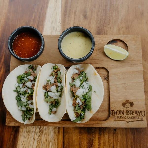 Three Don Bravo Mexican Grill tacos with meat, cilantro, and onions, served with two sauces and a lime wedge.