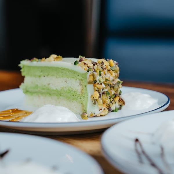 A slice of green pistachio cake with white frosting and chopped nuts on a white plate.