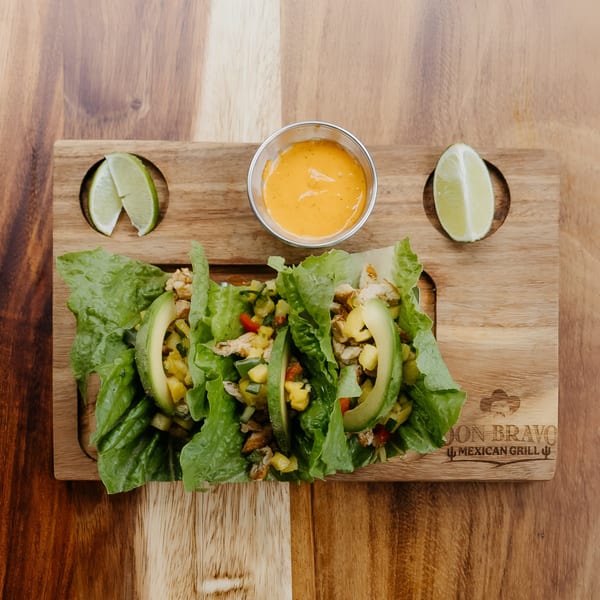 Three lettuce wraps with chicken, mango salsa, and avocado, served with lime wedges and sauce.
