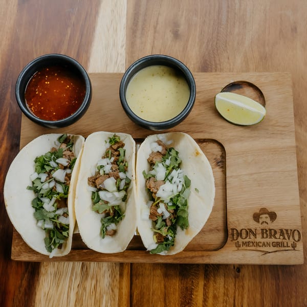 Three Don Bravo Mexican Grill tacos with meat, cilantro, and onions, served with two sauces and lime.