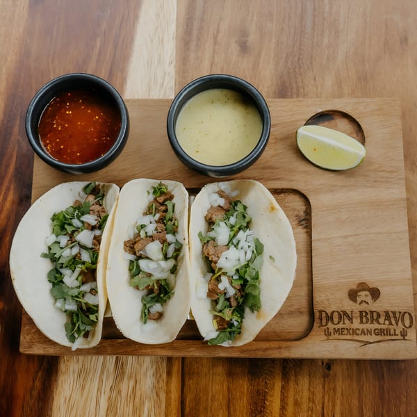 Three Don Bravo Mexican Grill tacos with meat, onions, and cilantro, served with two sauces and lime.