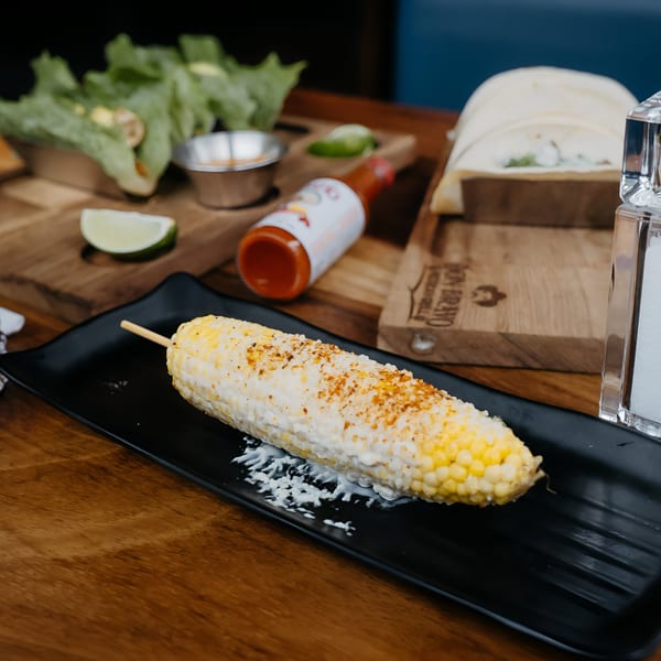 Elote, Mexican street corn, on a black plate with lime, hot sauce, and tortillas in the background.
