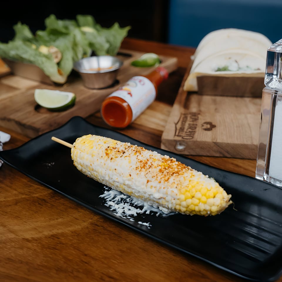 Elote, Mexican street corn, on a black plate with lime, hot sauce, and tortillas in the background.