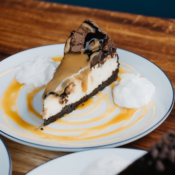 Slice of cheesecake with chocolate and caramel drizzle, and whipped cream on a white plate.