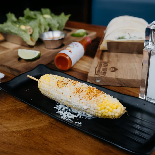 Elote, Mexican street corn, on a black plate with hot sauce, lime, and lettuce in the background.