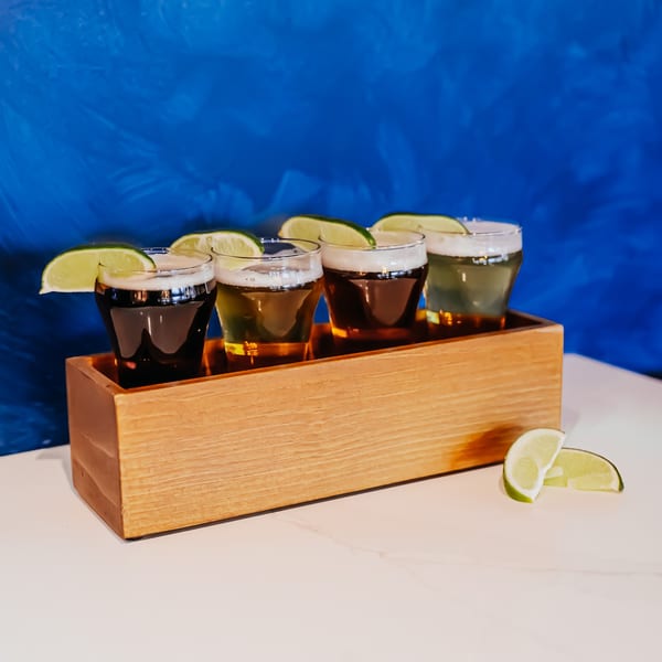 Four Don Bravo Mexican Grill beer samples in a wooden tray with lime wedges against a blue background.
