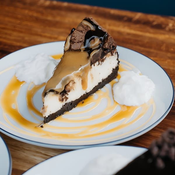 Slice of cheesecake with chocolate crust, caramel, chocolate drizzle, and whipped cream on a white plate.