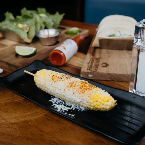 Elote, Mexican street corn, on a black plate with hot sauce, lime, and tacos in the background.