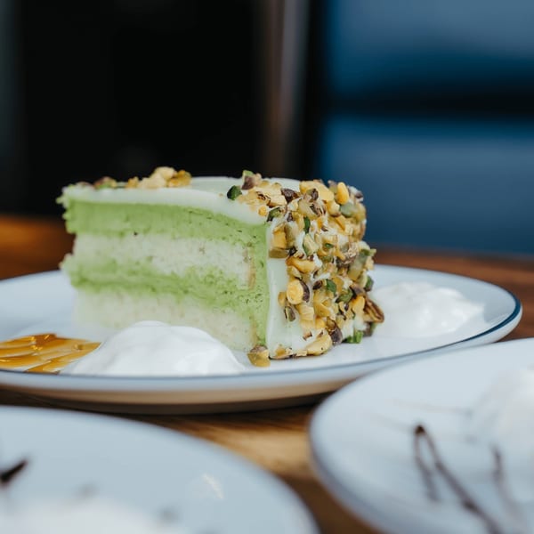 A slice of green pistachio cake with white frosting and nuts, served with cream and caramel.