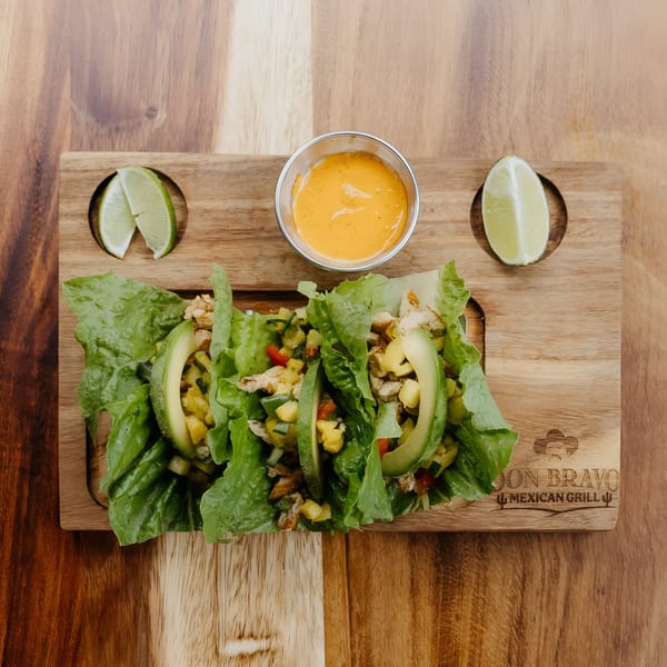 Three lettuce wraps with chicken, avocado, and salsa, served with lime wedges and sauce.