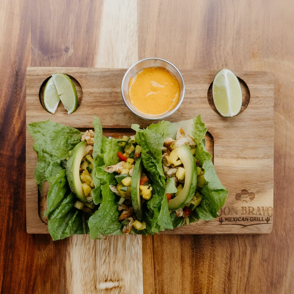 Three lettuce wraps with chicken, avocado, and salsa, served with lime wedges and sauce.