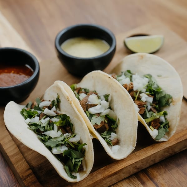 Three Don Bravo Mexican Grill tacos with meat, cilantro, and onions, with two sauces and a lime wedge.