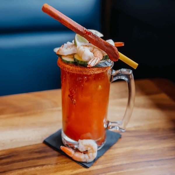 A michelada cocktail with a chili-rimmed mug, garnished with shrimp, cucumber, lime, and a tamarind candy straw.