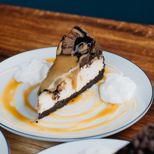 A slice of cheesecake with chocolate crust, caramel, and chocolate drizzle, served with whipped cream.