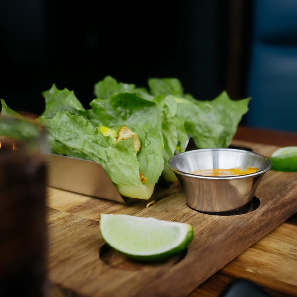Lettuce wraps with filling, a side of sauce, and lime wedges on a wooden board.