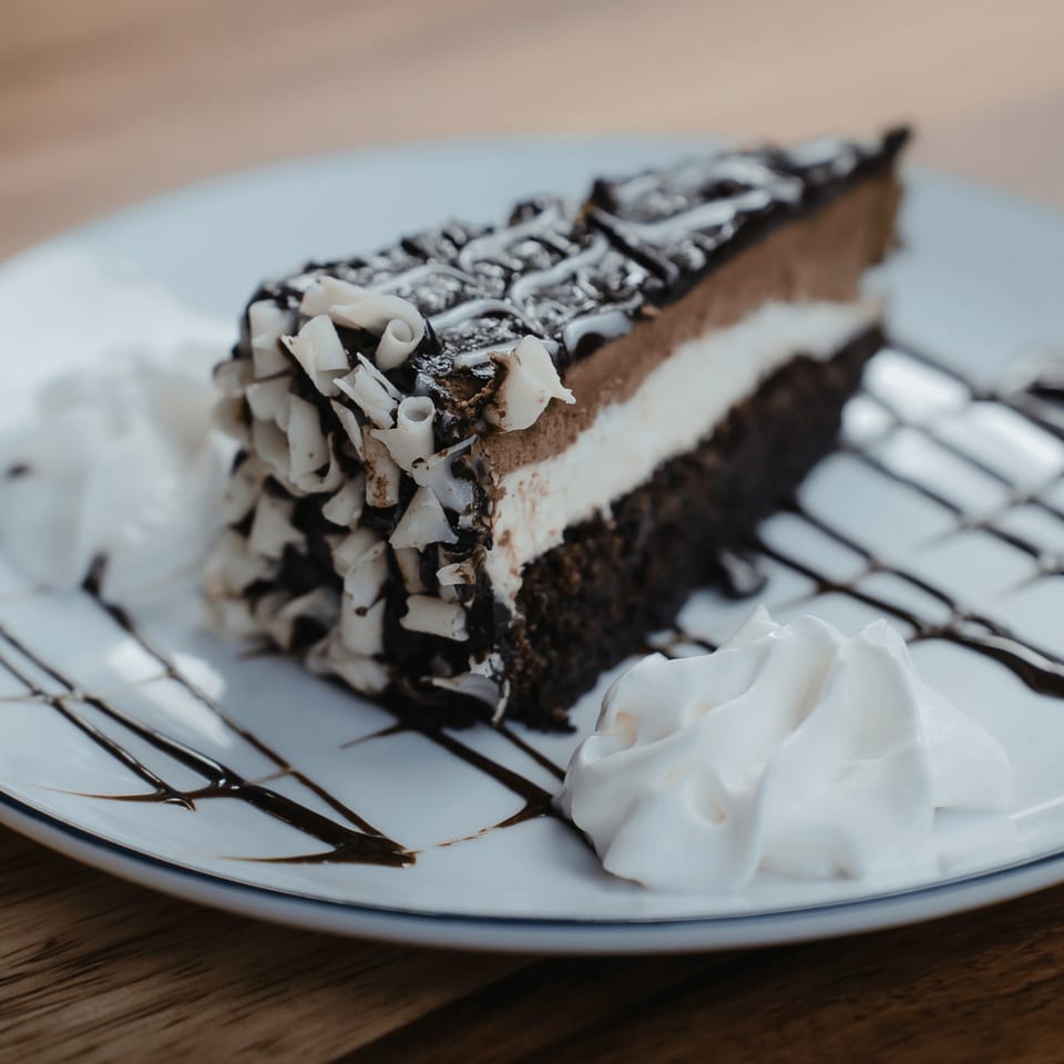 A slice of chocolate cake with white and milk chocolate layers, topped with chocolate shavings and whipped cream.