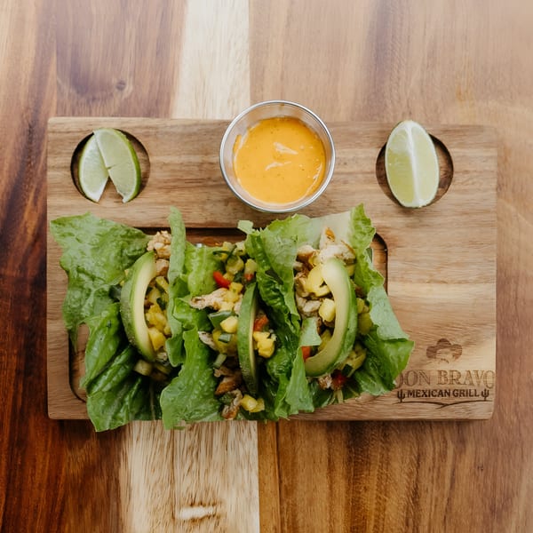 Three Don Bravo Mexican Grill lettuce wraps with avocado, salsa, and lime on a wooden board.